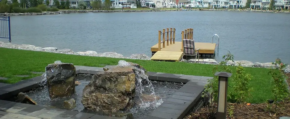 A custom stone rock fountain in a backyard overlooking a lake with a wooden swimming dock and ladder in Edmonton.