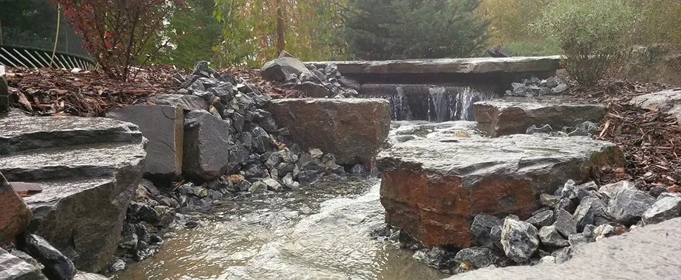A professionally constructed natural stone waterfall and stream feature with artistic rock placement and surrounding mulch in an Edmonton backyard.