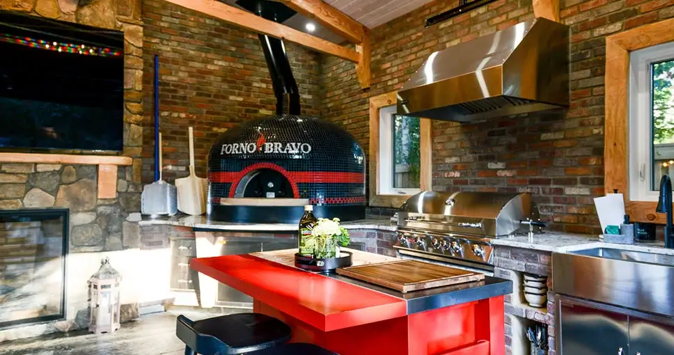 A luxury outdoor kitchen featuring a Forno Bravo pizza oven, stainless steel grill, and brick backsplash.