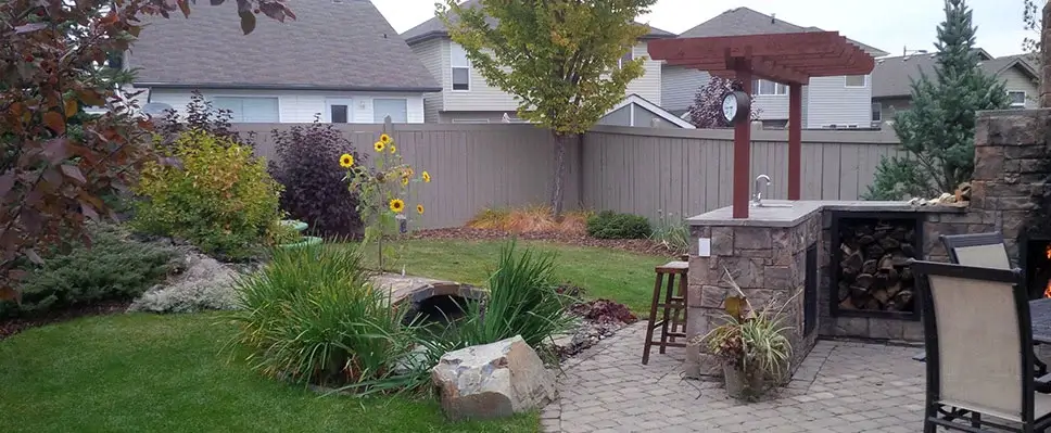 A stone-clad outdoor kitchen with a small red pergola, a wood storage nook, and a stone bridge over a garden pond in a residential Edmonton backyard.