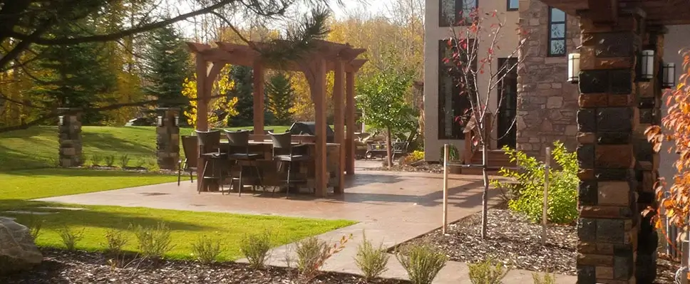 A luxury backyard featuring a stone-clad outdoor kitchen bar with high chairs under a custom timber pergola on a large patio in Edmonton.