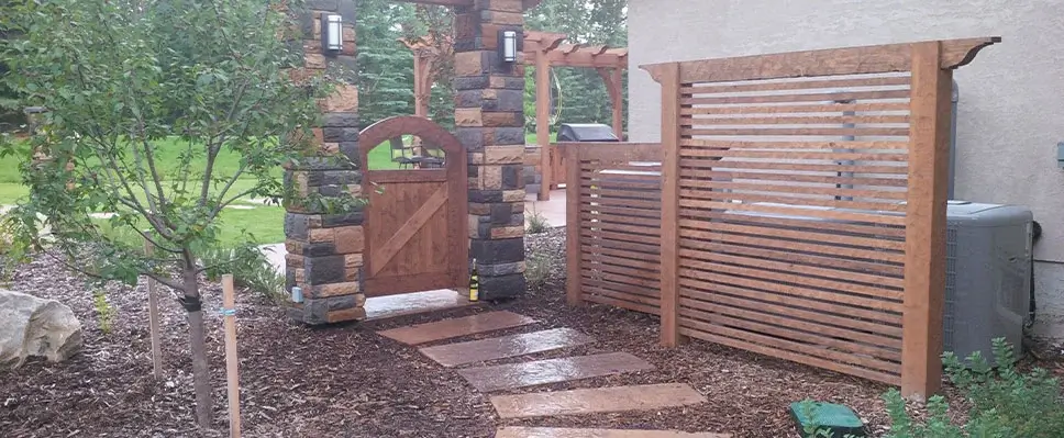 A custom-built wood garden gate and horizontal timber privacy screen integrated with stone pillars in an Edmonton backyard.