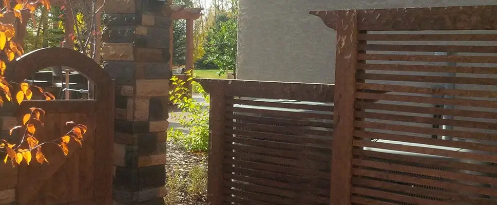A custom dark wood horizontal slat privacy screen next to a decorative stone pillar and arched wooden gate in an Edmonton backyard.