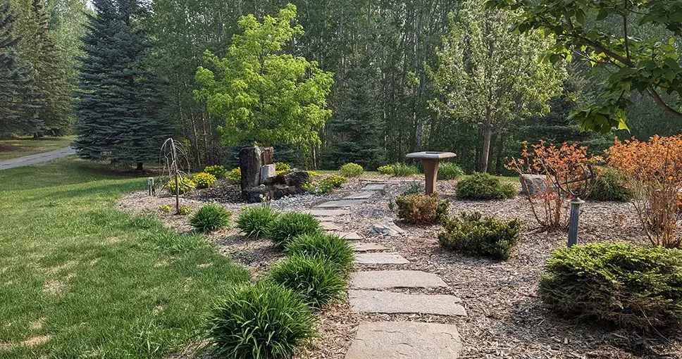 A scenic garden pathway made of stone slabs leading to a peaceful seating area with a birdbath and a decorative water feature, surrounded by lush greenery.