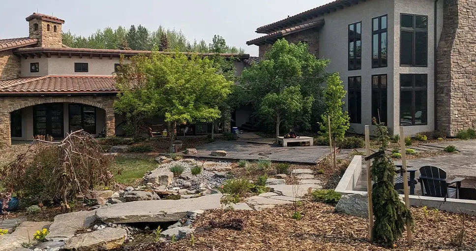 A Mediterranean-style home with a natural stone pathway, a dry creek bed, and a wooden deck surrounded by lush greenery.