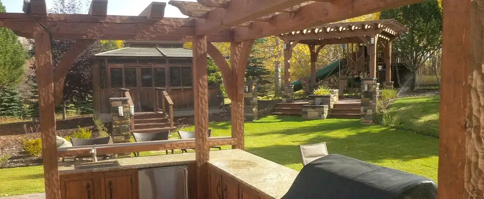 View from a custom outdoor kitchen looking out toward a wooden gazebo, multiple pergolas on stone decks, and a children's playset in a large Edmonton backyard.