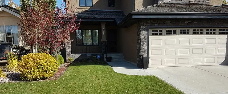 A modern residential front yard in Edmonton featuring a clean concrete driveway, stone-clad exterior pillars, a lush green lawn, and decorative yellow and red shrubs.