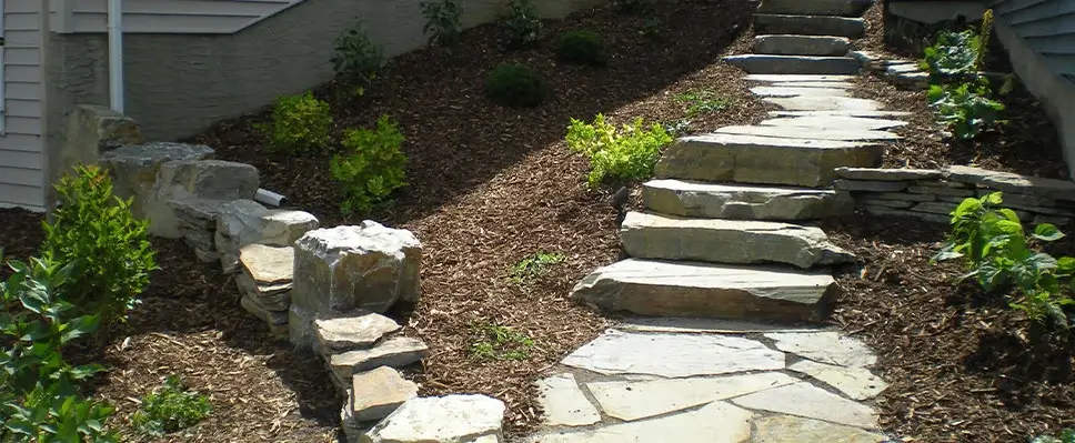 A winding natural flagstone walkway and stone retaining wall built on a sloped garden bed with dark mulch and small green plants in Edmonton.