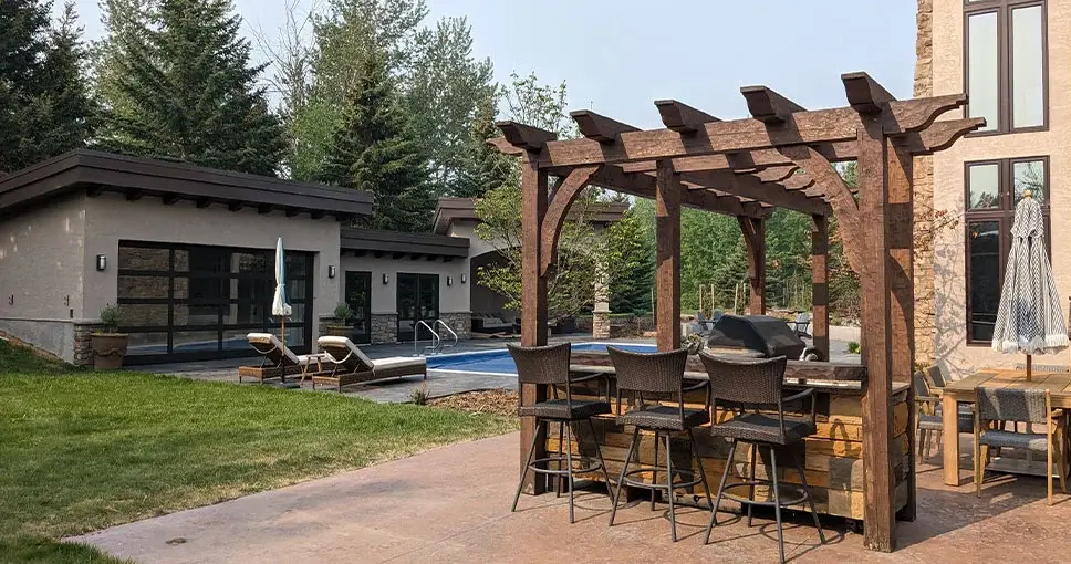 A large dark wood pergola over an outdoor bar and kitchen area next to a swimming pool.