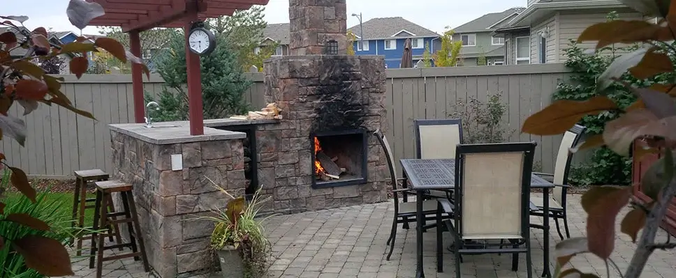 A custom stone outdoor fireplace and kitchen island with a dining table set on a paver patio in an Edmonton backyard.