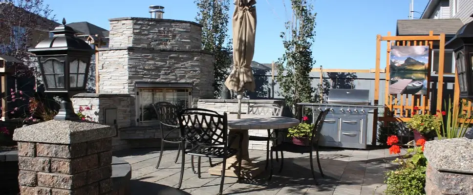 A stone paver patio in Edmonton featuring a large outdoor stone fireplace, a stainless steel gas grill, and a circular dining table with black wrought iron chairs.
