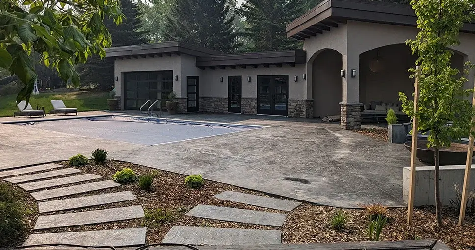 A modern outdoor poolside area with a stone pathway, landscaped greenery, and a covered lounge space.