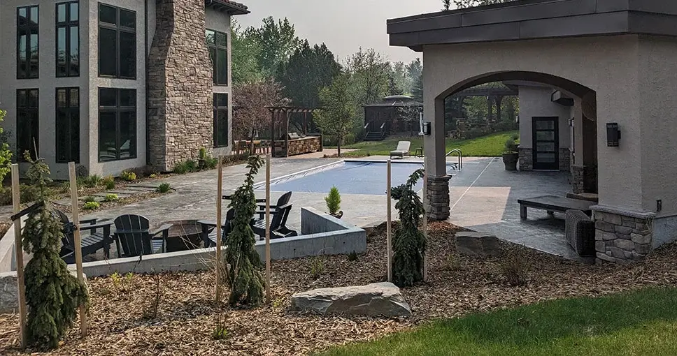 A luxurious backyard with a modern swimming pool, fire pit seating area, and a covered patio surrounded by lush landscaping.