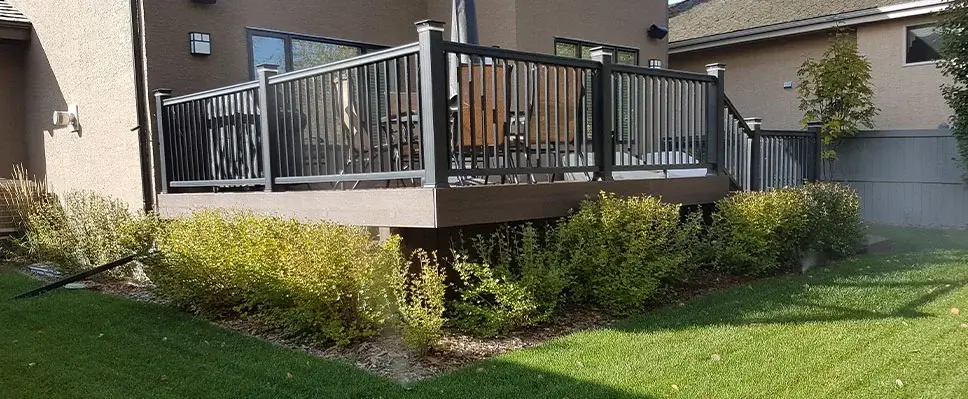 A large, raised brown composite deck with black aluminum railings and post cap lights, surrounded by manicured shrubs and a green lawn in Edmonton.