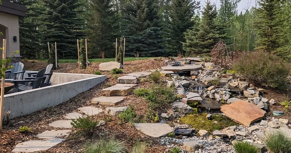 A scenic landscape featuring a stone pathway alongside a cascading water feature, surrounded by lush greenery and outdoor seating.