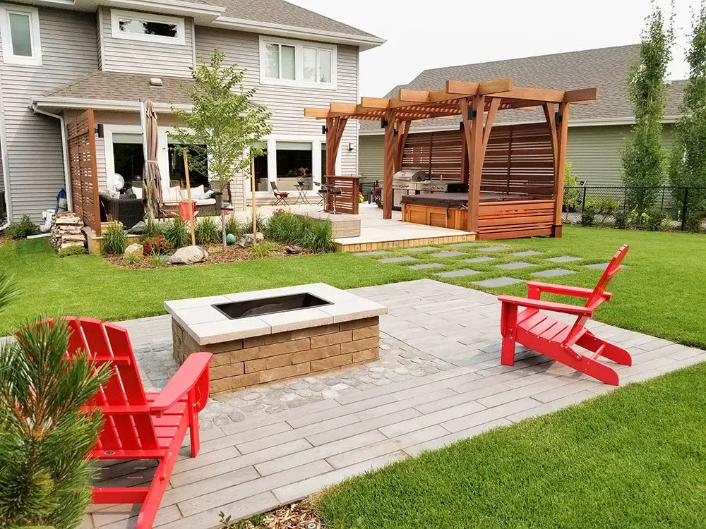 Backyard landscaping in Edmonton with fire pit, red Adirondack chairs, wooden pergola-covered hot tub, & raised wooden deck.