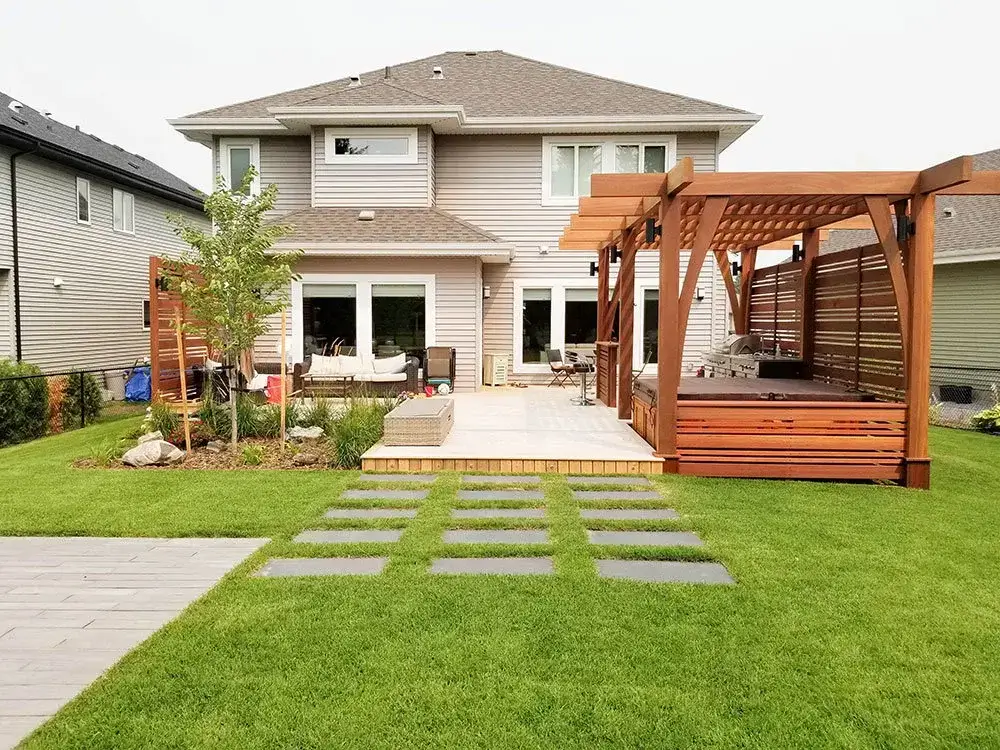 Backyard landscaping in Edmonton with pergola-covered hot tub, wooden deck, stone pathway, and landscaped garden bed.