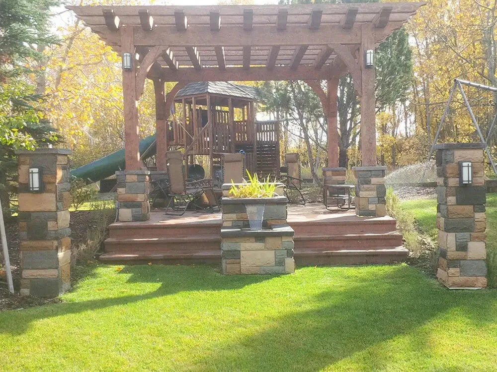 Family-friendly backyard landscaping in Edmonton with wooden pergola, patio seating, play structure, and autumn trees.