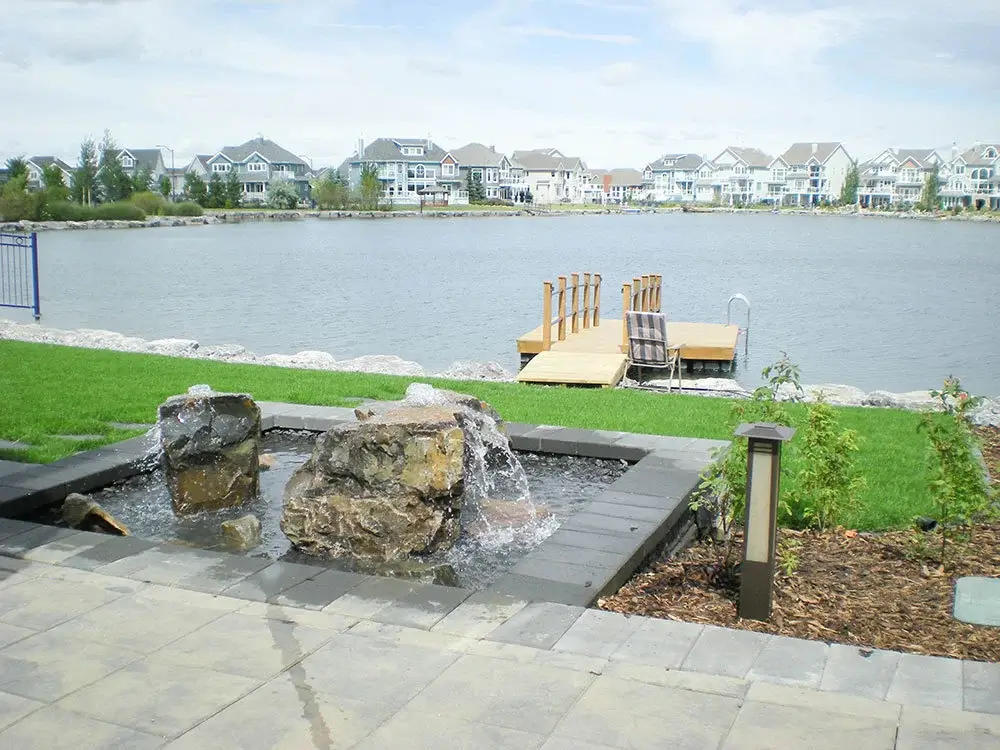 Lakeside Edmonton landscape with water feature, stone patio, garden bed, and wooden dock.