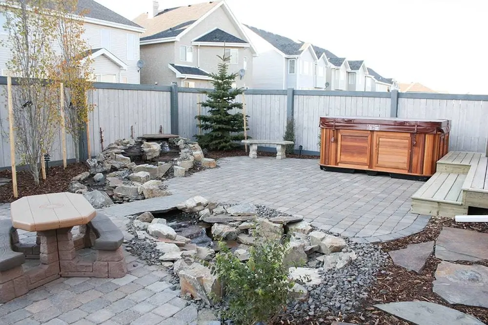 Backyard with stone patio, waterfall feature, hot tub, and privacy fencing showcasing landscape construction Edmonton.