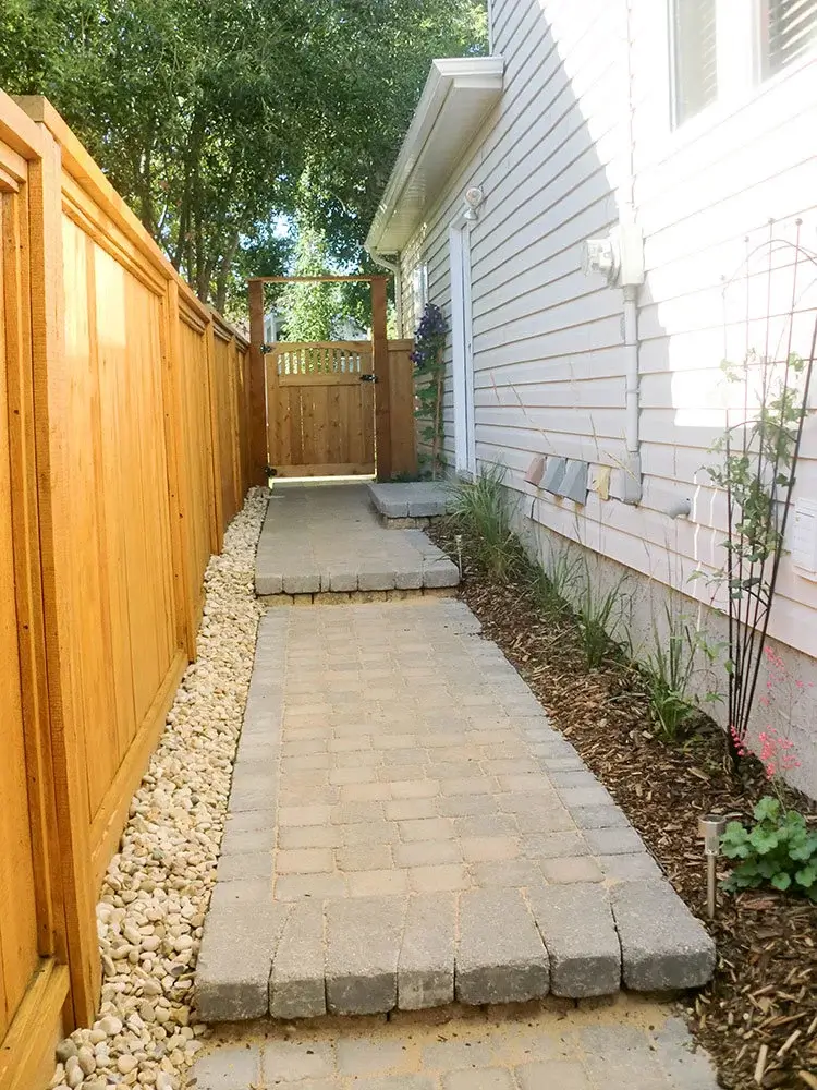 Side yard Edmonton landscape with stone pathway, wooden fence, garden bed, & flowering trellis.