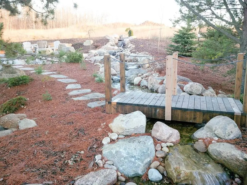 A peaceful garden with a wooden bridge, stream, and fire pit—part of custom landscaping construction in Edmonton