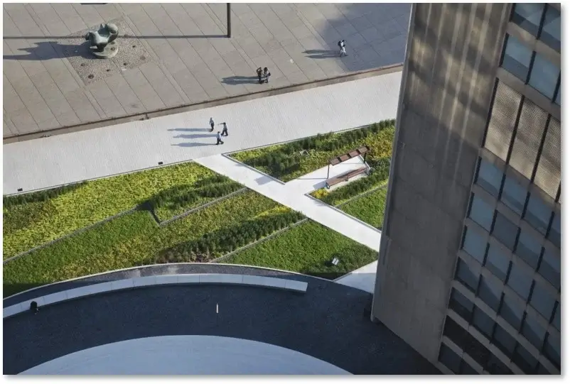 An aerial top-down view of a modern urban plaza featuring geometric green roof sections, white pedestrian walkways, and people walking near a tall building.