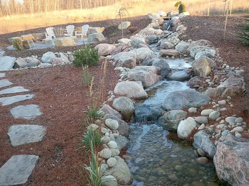 A long natural stone creek water feature leading toward a rustic rock fire pit with Adirondack chairs.