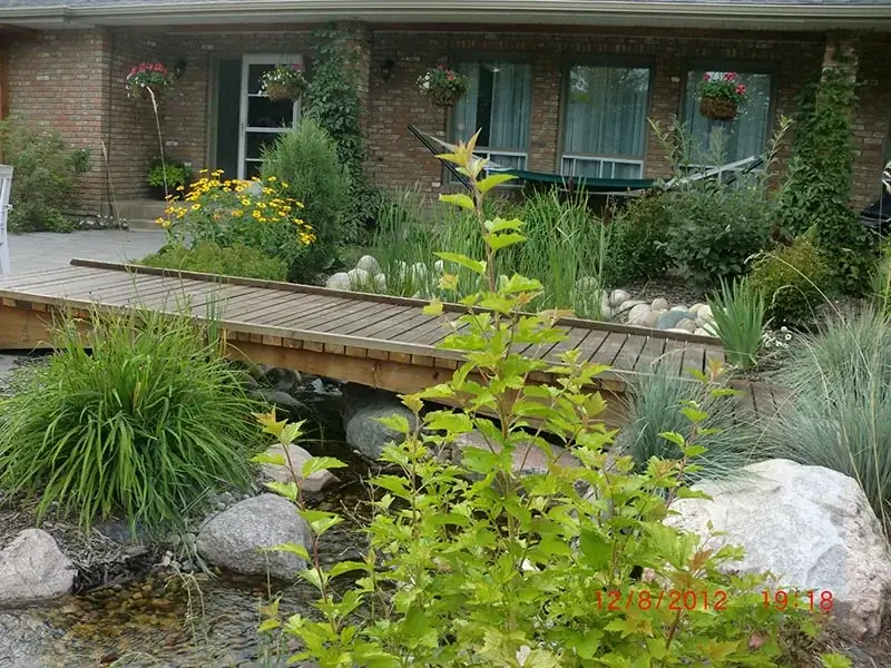 A small wooden footbridge over a rocky stream in a lush residential garden with flowers and a brick house.