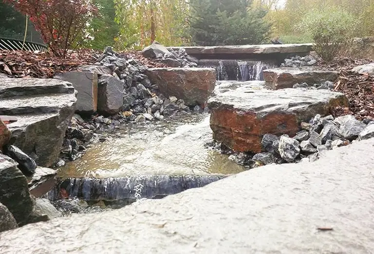 A recirculating backyard stream flowing over large natural boulders and dark mulch.