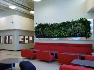 A long, horizontal living wall with diverse green plants in a silver metal frame above red lounge seating in a large commercial building atrium.