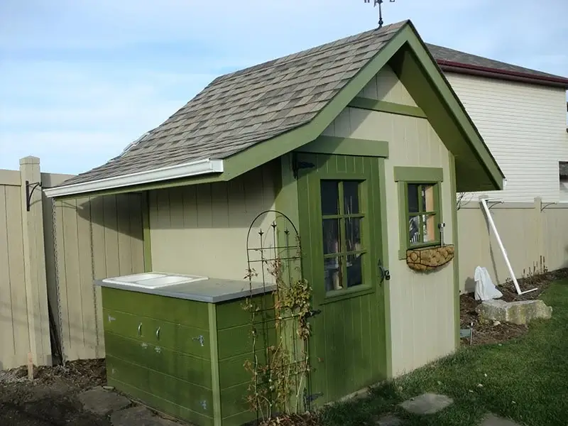 A small tan garden shed with green trim, a shingled roof, and an integrated outdoor double sink.