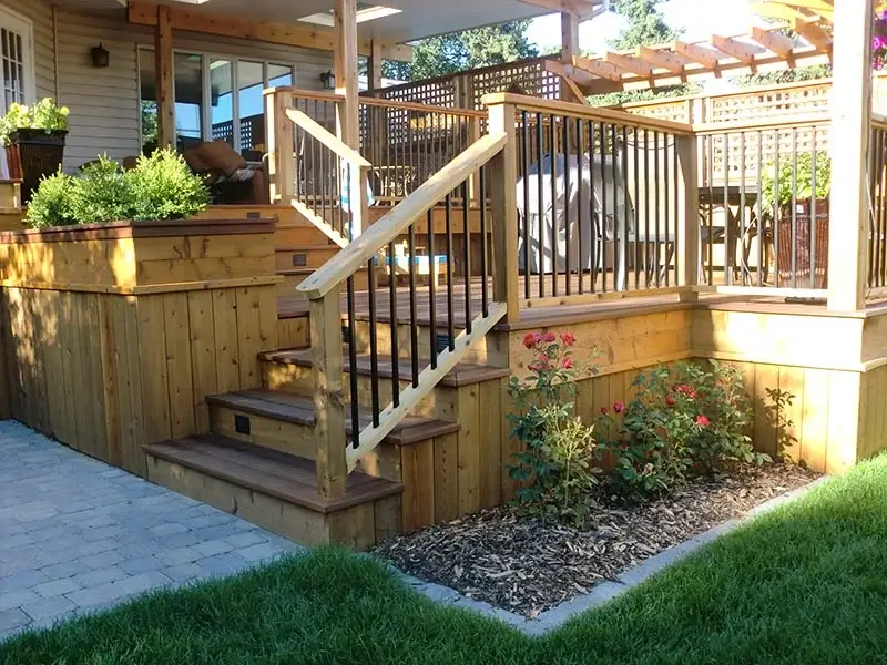 A multi-level wooden deck with black metal railings, built-in planters, and integrated step lighting next to a stone patio.