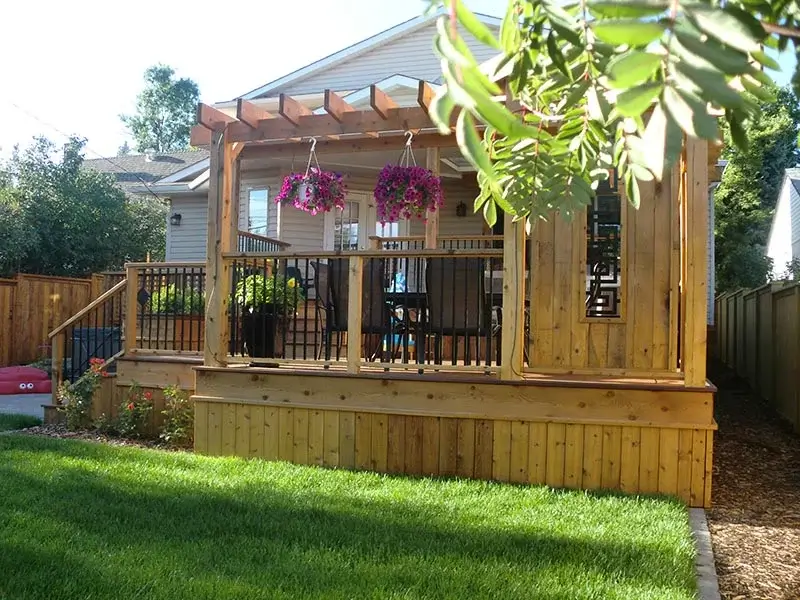 A multi-level wooden deck featuring a pergola, hanging flower baskets, and a dining area with black railings.