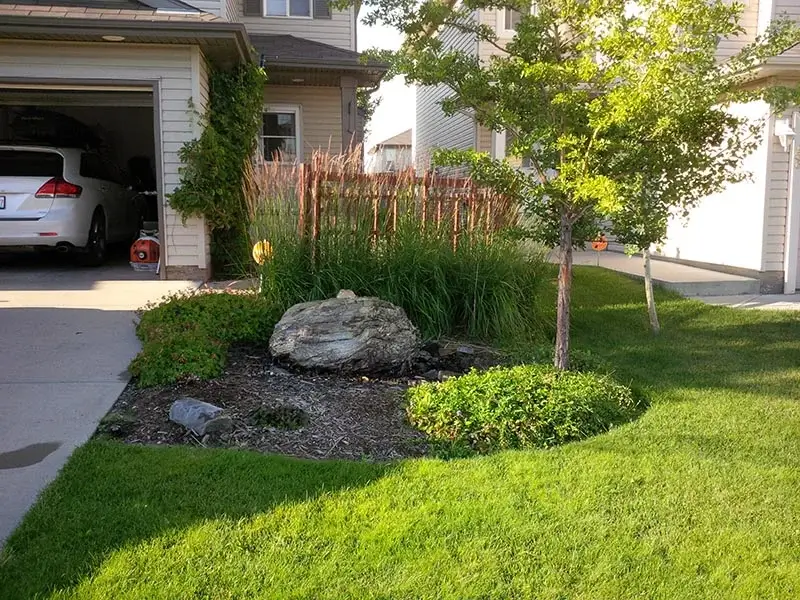 A natural stone water fountain in a front yard garden bed surrounded by ornamental grasses and a green lawn.
