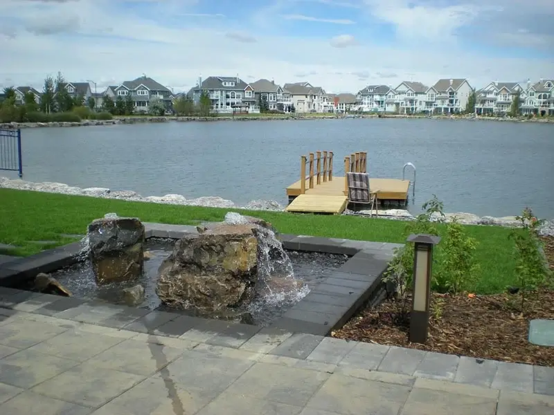 A backyard stone patio featuring a rock waterfall fountain overlooking a lake and private dock.