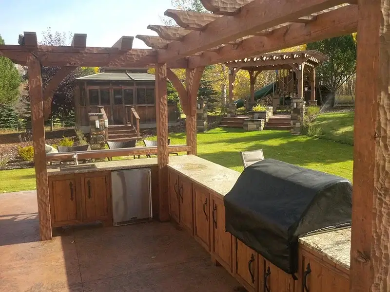 A custom outdoor kitchen with granite countertops, wooden cabinets, and a heavy timber pergola overlooking a manicured lawn.