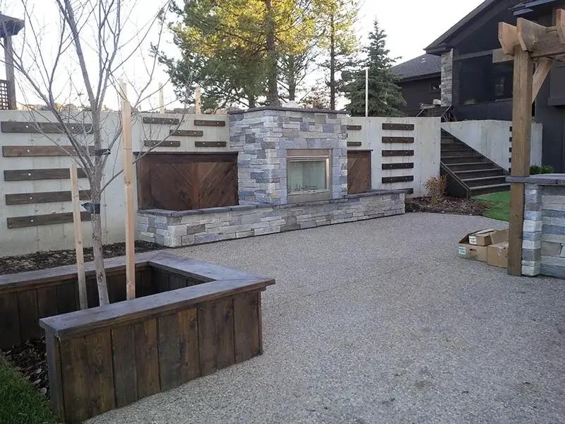 A modern outdoor stone fireplace on an aggregate patio with wooden privacy wall accents and a built-in planter.