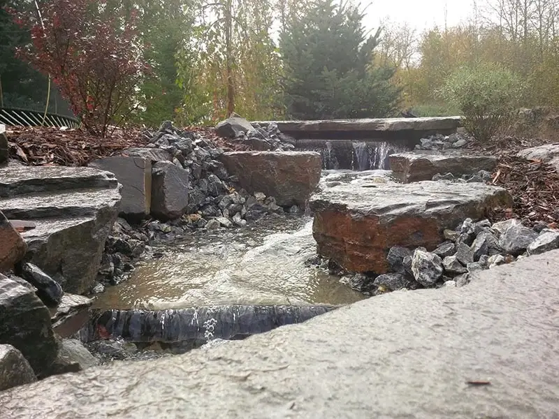 A multi-tiered natural rock waterfall cascading into a stream with dark mulch and evergreens in the background.
