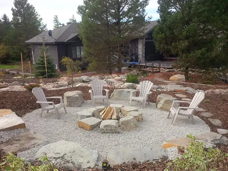 A circular gravel fire pit area with a ring of large rocks, Adirondack chairs, and surrounding forest landscape.