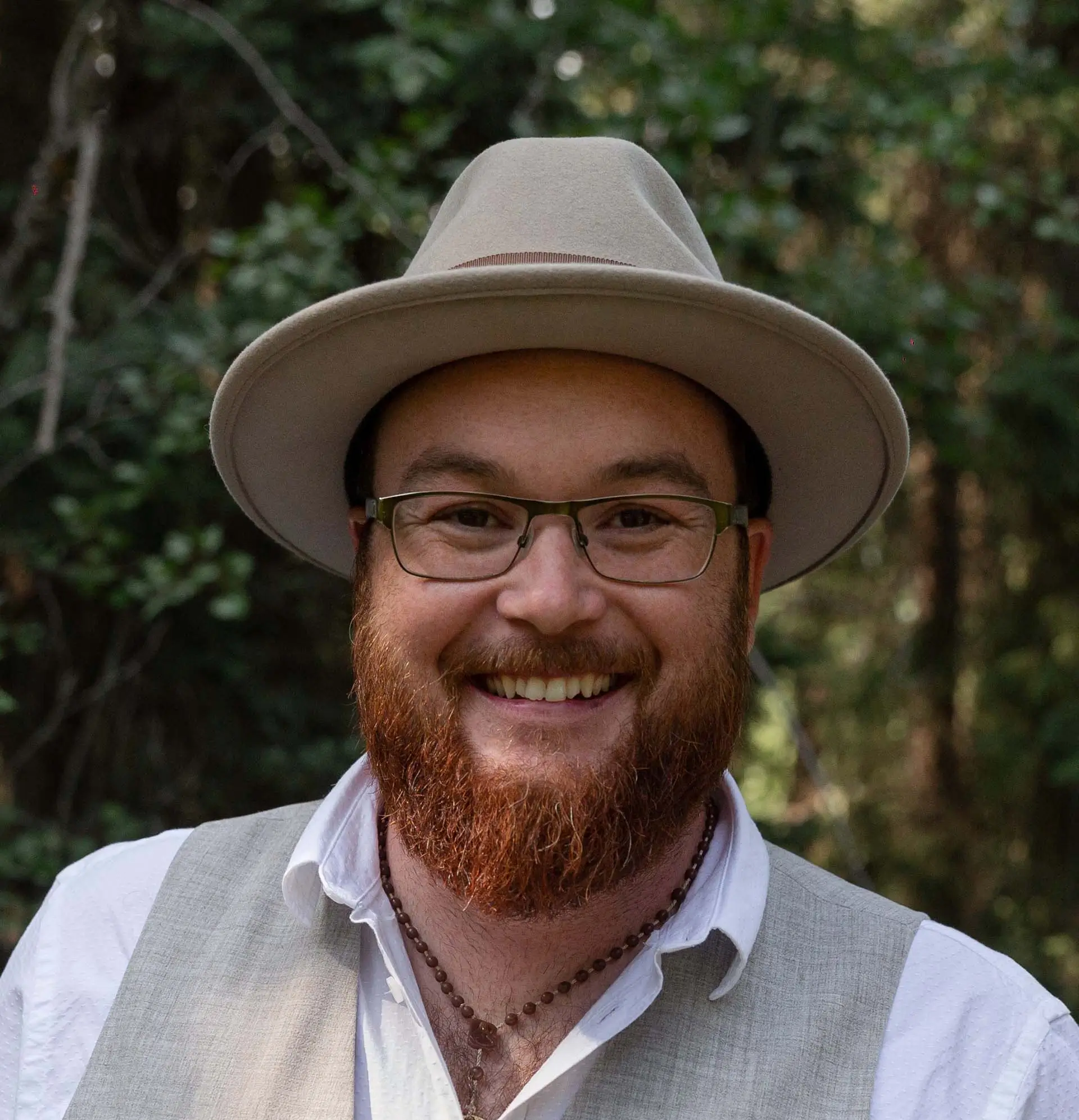 A professional headshot of a smiling man with a red beard wearing a tan fedora, glasses, and a grey vest against a soft forest background.