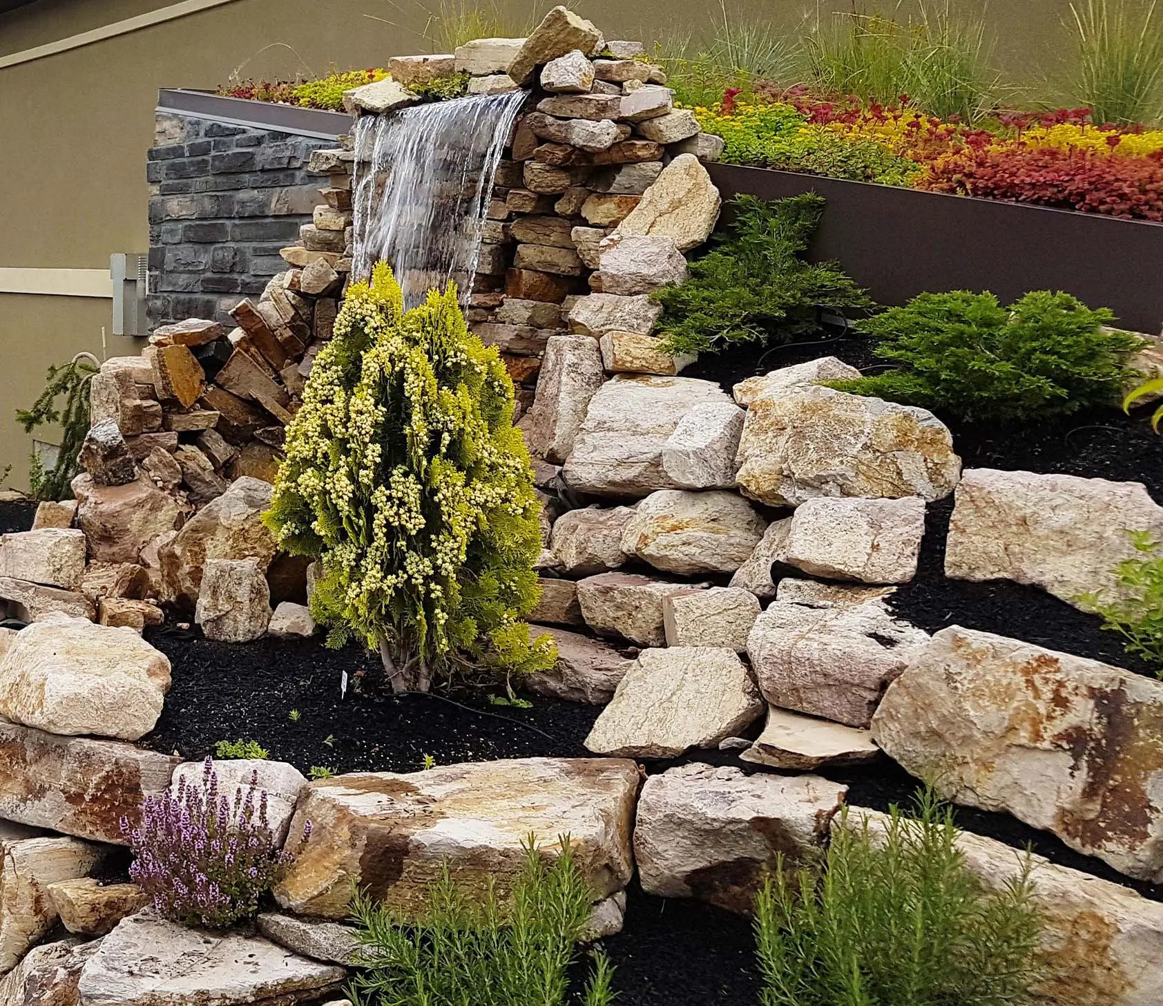 A custom natural stone waterfall built into a tiered rock retaining wall with ornamental shrubs and a colorful garden bed.