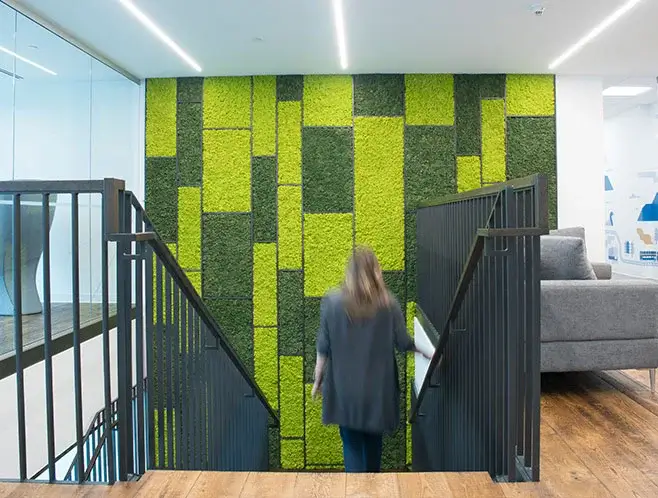 Large two-tone (lime and dark green) geometric preserved moss wall installation spanning multiple floors behind a modern office staircase with metal railings.