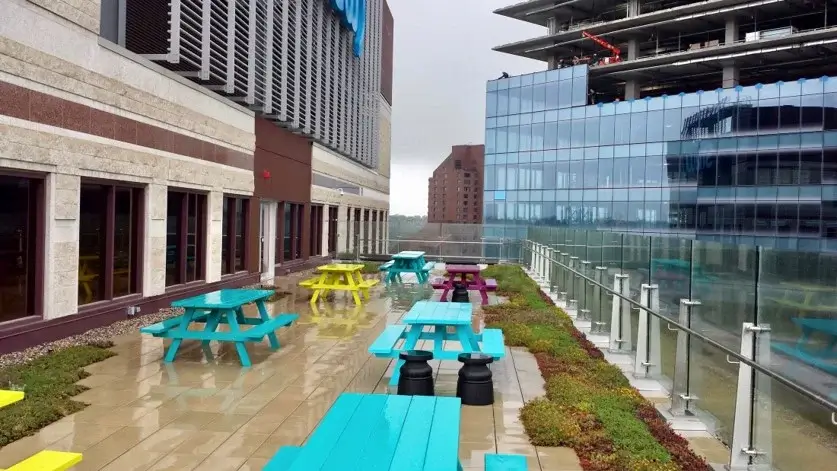 A modern rooftop patio with colorful picnic tables, glass railings, and integrated green roof borders.