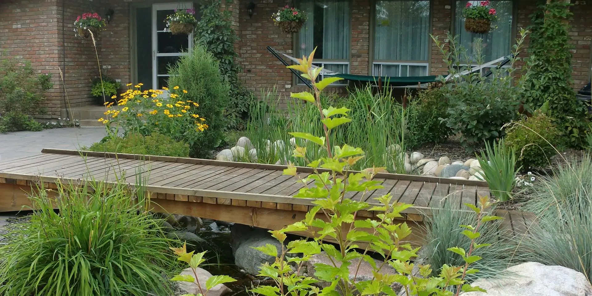 Beautiful landscape design featuring a wooden bridge over a pond, lush greenery, and vibrant flowers in a well-maintained garden.