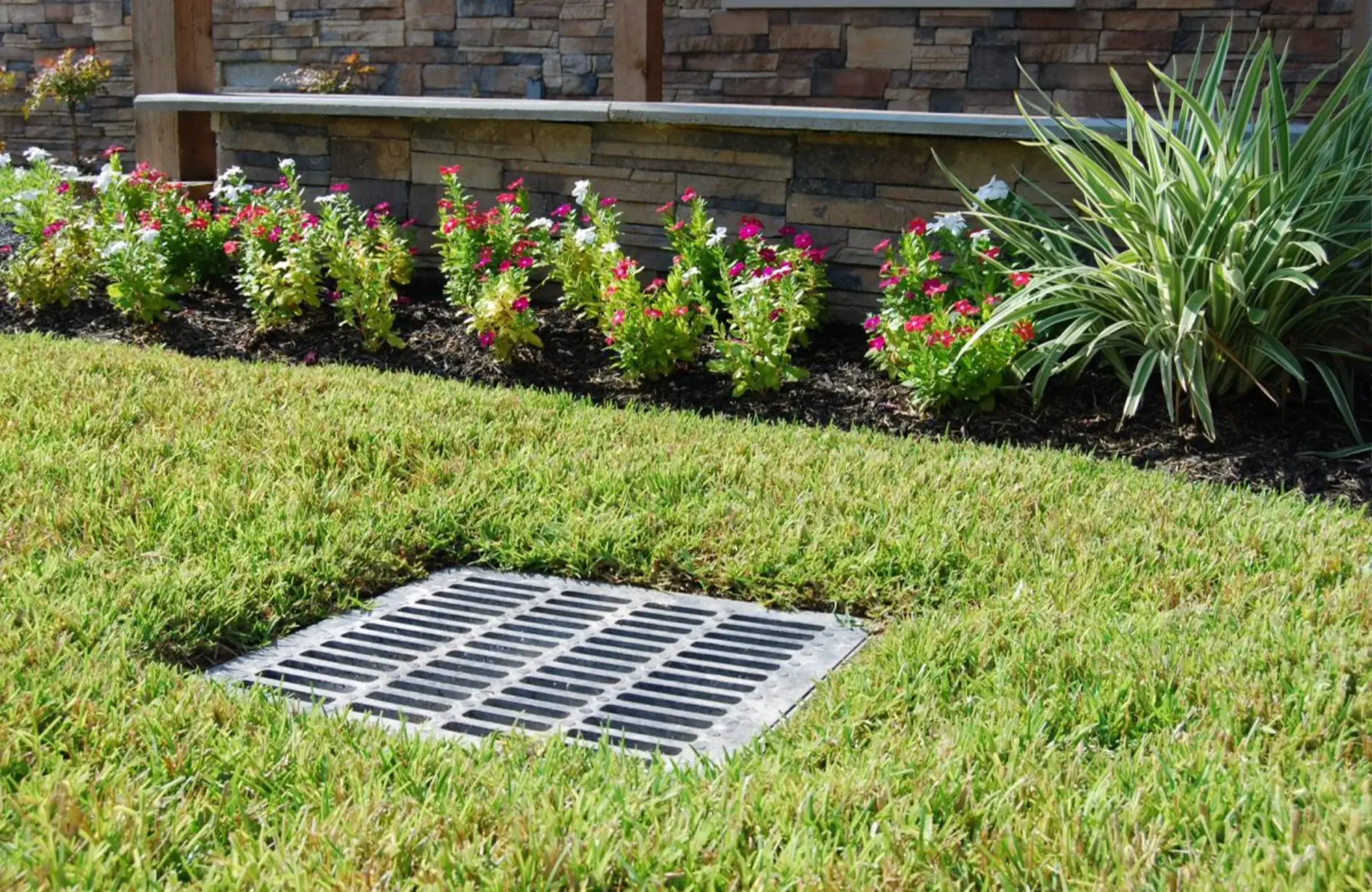 Yard drainage solution with a grate in grassy area, surrounded by colorful flowers and a stone wall.