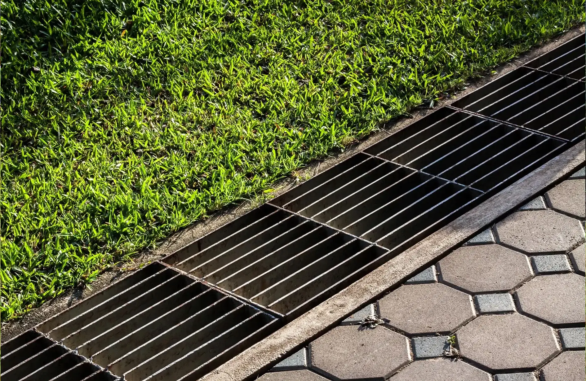 Yard drainage solution with a long grate between grassy area and patterned pavement.